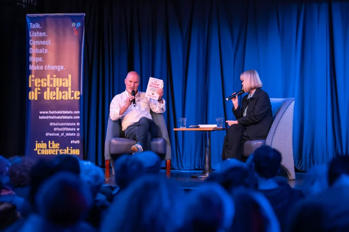 A photo of two people sitting onstage during a talk event, one is holding up a book titled 'The Good Society' and next to them is a big banner with "Festival of Debate" on it