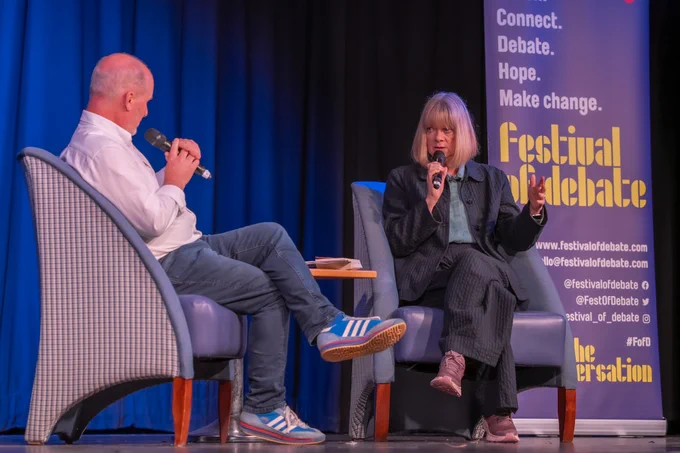 A photo of two people sitting on a stage in conversation, holding microphones. Behind them is a big banner with "Festival of Debate" on it