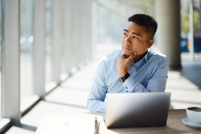 Man in a light blue shirt sitting at a desk with a laptop, resting his chin on his hand, looking thoughtfully out a large window.