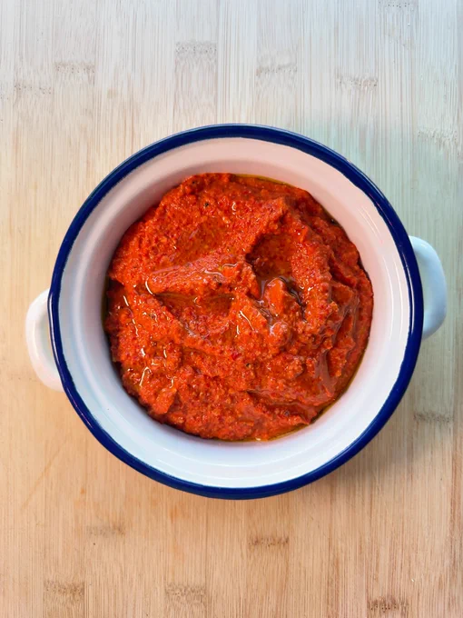 Cuenco blanco con muhammara de color rojo intenso, preparada con pimientos del piquillo, nueces, ajo, lima, especias y aceite de oliva virgen extra. La crema aparece rematada con un chorro de aceite por encima y servida sobre una mesa, lista para mojar pan o acompañar otros platos.