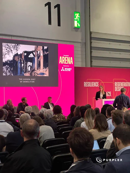 This image captures a presentation titled 'The Hidden Cost of Demolition' at a conference or expo. The setting includes seated attendees and presenters with a focus on discussions around sustainability and construction.