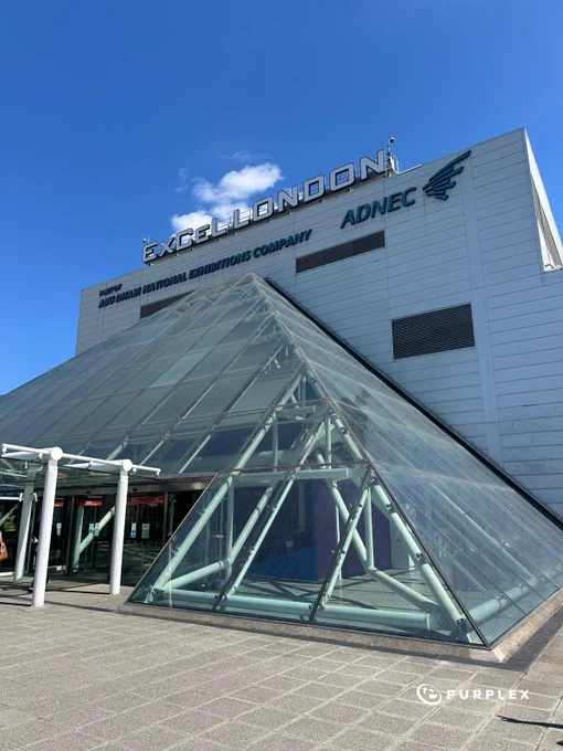 A bright and clear day view of the ExCeL London exhibition center with its modern glass pyramid structure and the ADNEC signage prominently displayed.