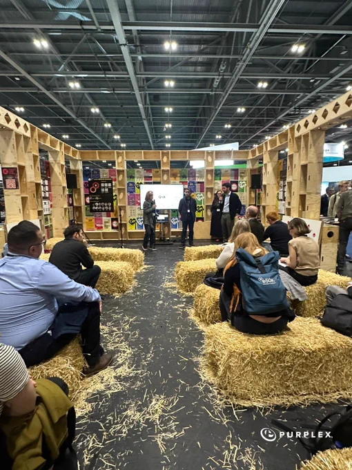 Visitors seated on hay bales listening to a panel discussion in a creatively designed exhibit area. The setting exudes an informal and engaging atmosphere.