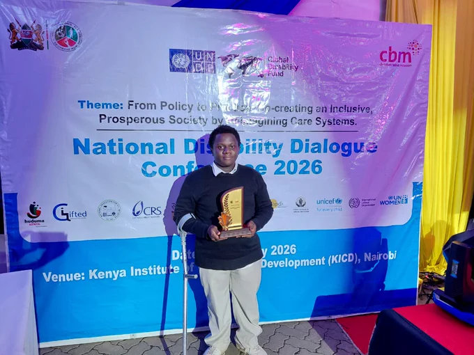 A person stands on a red carpet holding an award plaque during the National Disability Dialogue Conference 2026 at the Kenya Institute of Curriculum Development (KICD) in Nairobi. The conference backdrop features logos from organizations including UNDP, Global Disability Fund, CBM, UNICEF, and UN Women, with the theme focused on creating an inclusive and prosperous society through reimagining care systems. The person is smiling and posing with a mobility aid beside them.