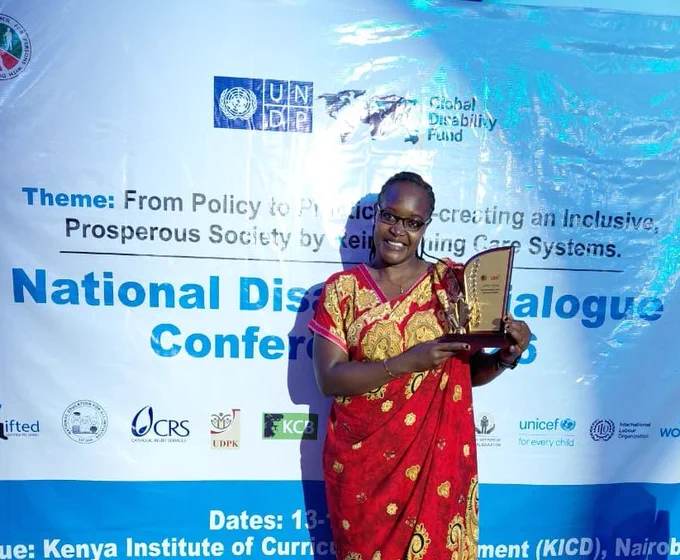 A woman wearing a red and gold patterned dress stands smiling while holding an award plaque at the National Disability Dialogue Conference 2026. She is posing in front of a conference backdrop displaying the event theme on creating an inclusive and prosperous society through reimagining care systems, along with logos of organizations including UNDP, UDPK, Global Disability Fund, UNICEF, CRS, KCB, and UN Women. The event took place at the Kenya Institute of Curriculum Development (KICD) in Nairobi.