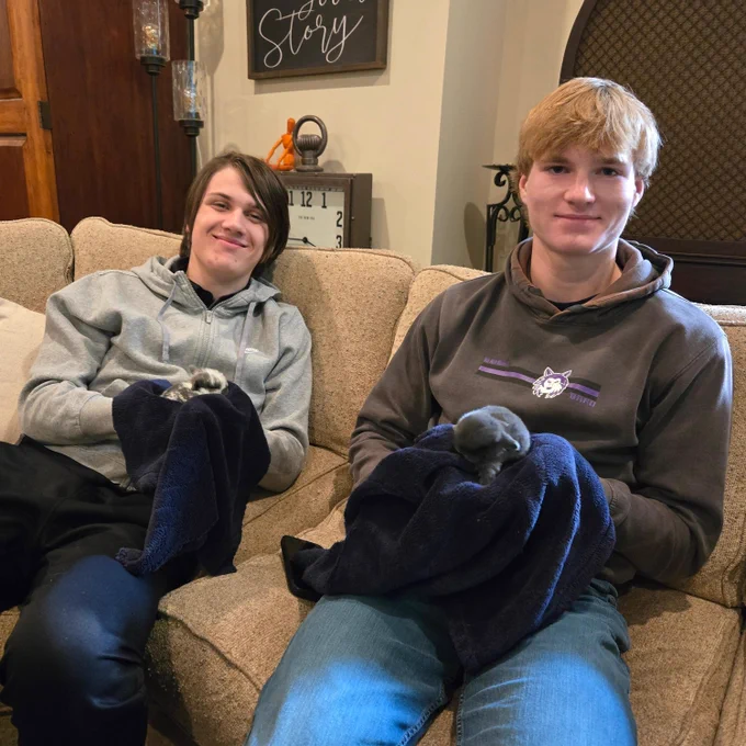 Two people sitting on a couch, smiling and wearing casual clothing, as they hold baby bunnies in towels.