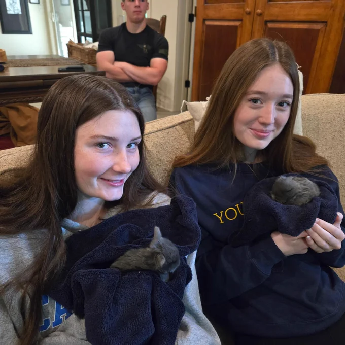 Two young women in black sweatshirts smile and pose for the camera as they hold baby bunnies, with a man in the background.