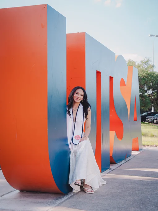 A person in a graduation outfit smiles while leaning against large, colorful letters spelling "UTSA" outdoors. The background includes trees and a clear sky.