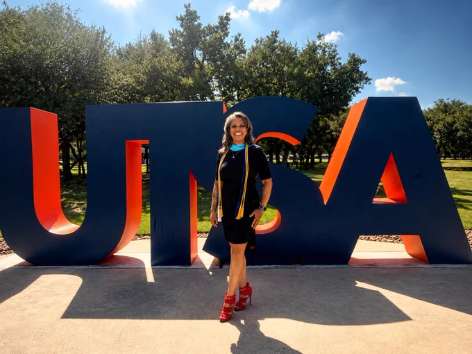 A person is standing in front of large, colorful letters spelling "UTSA" outside. They are wearing a black outfit with a colorful sash and red shoes. The background features green trees and a clear blue sky.