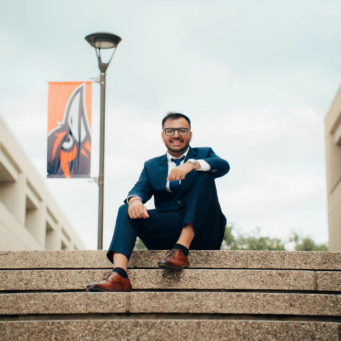A person in a blue suit sits on outdoor steps, smiling. A lamppost and a banner with the UTSA Roadrunners logo are visible in the background.