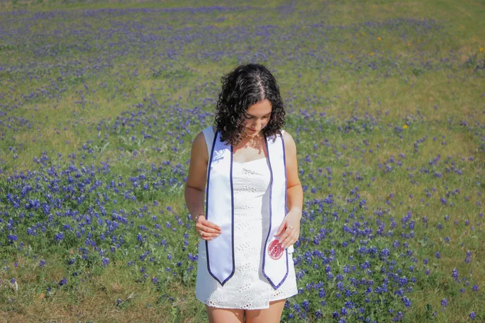A person wearing a white outfit and a graduation stole looks down while standing in a field of purple flowers.