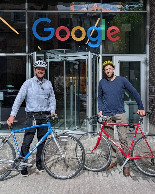 Two Googlers wearing helmets stand with their bicycles in front of a Google office building.
