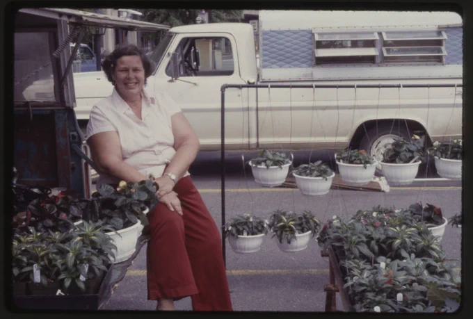 An unidentified, smiling woman sits outside in a farmer's market booth. She is surrounded by a display of potted, live plants while a white pickup truck is located in the background. 