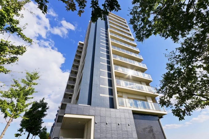 Modern high-rise apartment building with balconies under a bright blue sky framed by green trees.