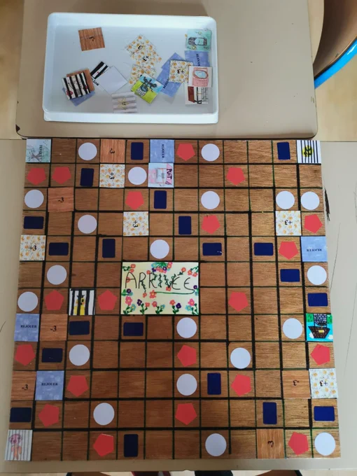Plateau de jeu fabriqué par des élèves, composé d’une grille en bois avec des cases colorées et illustrées (bonus, malus, actions), accompagné de cartes dessinées à la main posées dans un plateau au-dessus.
