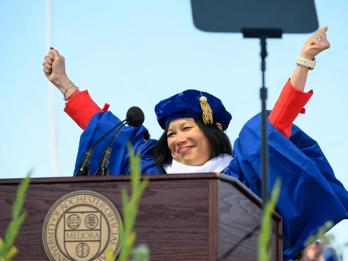 Jeannine Shao Collins, wearing a commencement cap and gown, raises her hands above her head in celebration from behind the podium. 