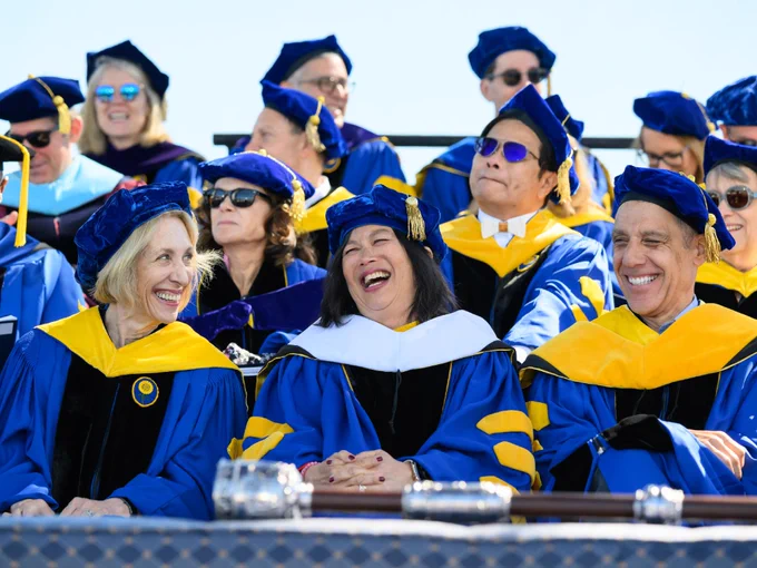 Jeannine Shao Collins laughs on stage as she sits among members of the platform party all dressed in commencement regalia. 