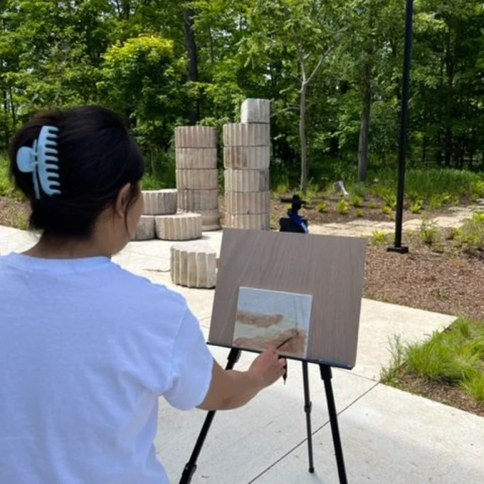 Person painting a landscape outdoors with trees and stacked stone columns in the background on a sunny day.