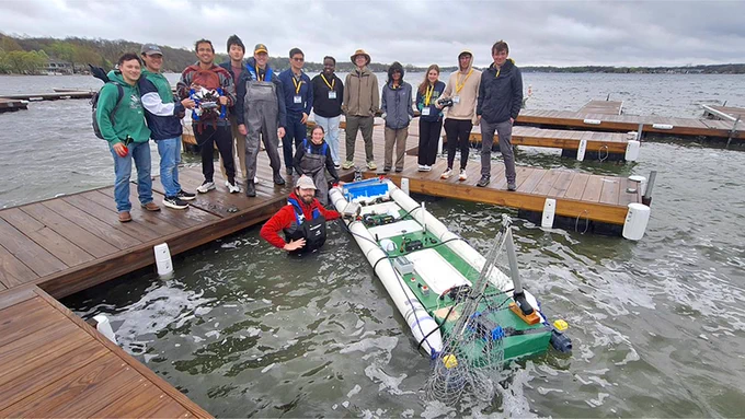 Team members stand on a dock beside Notre Dame’s AI modular boat during the AIMM ICC 2026 competition. One team member is in the water next to the vessel, which features a dynamic water-ballast system.