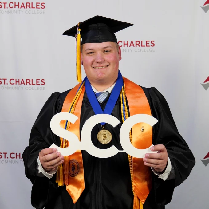 A person in graduation attire holding a decorative "SCC" sign. They are wearing a cap and gown with orange and blue stoles. The background features a "St. Charles Community College" logo.