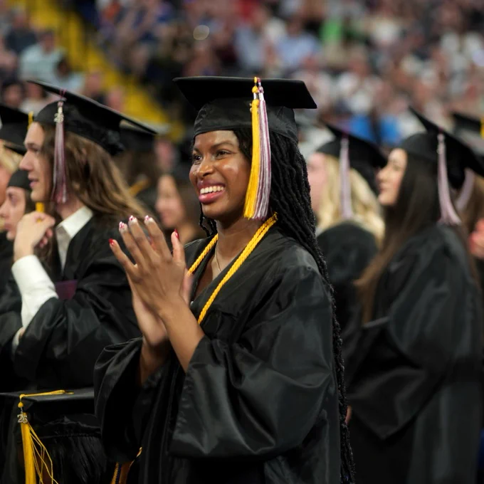 A person in a graduation cap and gown stands clapping among other graduates. A crowd is visible in the background, celebrating the event.