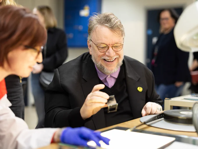 A man smiling, looking down at a film reel. A person on the left side of the image, wearing plastic gloves, is feeding the reel of the viewing desk. 