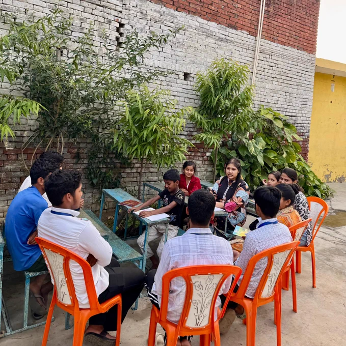A group of children, teenagers, and young adults sit together in a circle outdoors at Bansa Library, engaged in a book discussion. Some are seated on bright orange chairs while others sit on benches around a table with books and papers. The gathering takes place beside a brick wall lined with green plants, creating an open and informal community learning space where members from different age groups are listening and sharing ideas together.