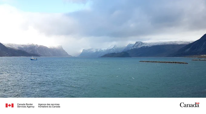Vast body of water framed by towering mountains with snow on their tops, under thick clouds and a distant boat.