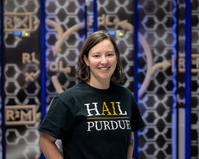 Woman wearing a black T-shirt with 'HAIL PURDUE' printed in white and gold letters standing against a geometric patterned background.