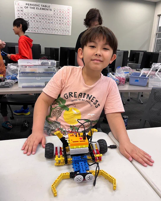 Young boy proudly displays a colorful LEGO robot built with wheels and sensors on a classroom table. Other kids and computers in background at Chattanooga State. 