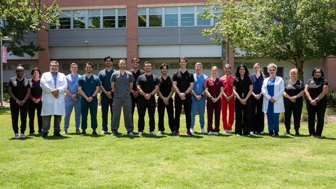 A large group of people stands in a line outdoors on a grassy courtyard area in front of a brick building. They wear a mix of medical scrubs in various colors and white lab coats, posing together with their hands crossed.