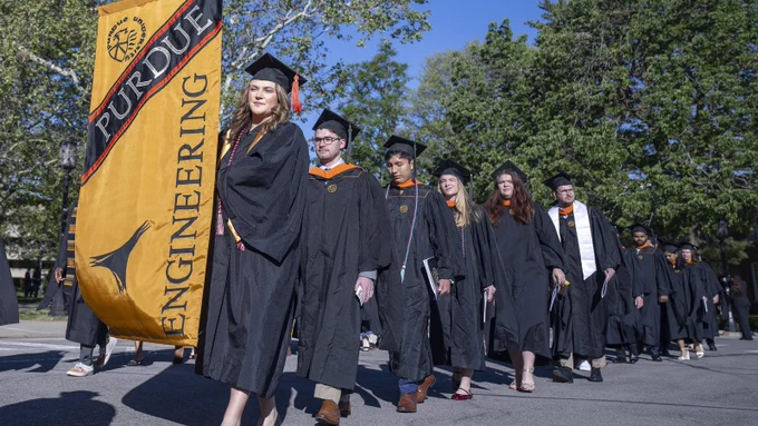 A long line of Purdue Engineering graduates in black gowns march in the commencement procession under blue skies, led by a graduate carrying a large gold and black Purdue Engineering banner.