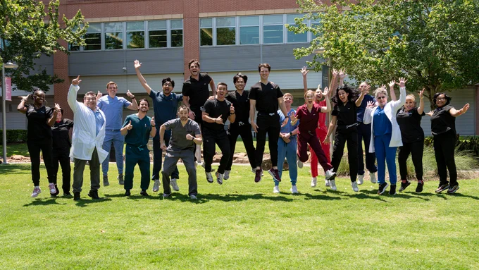 A large group of people wearing medical scrubs and white lab coats jumps in the air together on a grassy courtyard in front of a brick building.