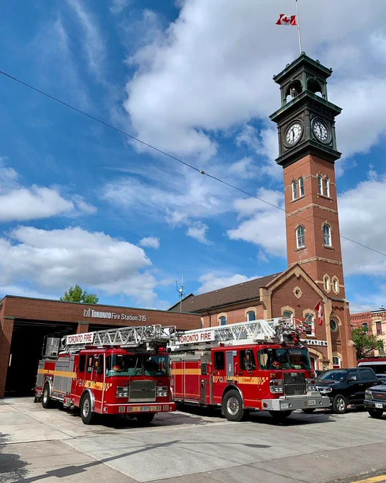 Two red Toronto Fire trucks parked outside Fire Station 315 with a tall clock tower flying a Canadian flag against a partly cloudy sky.