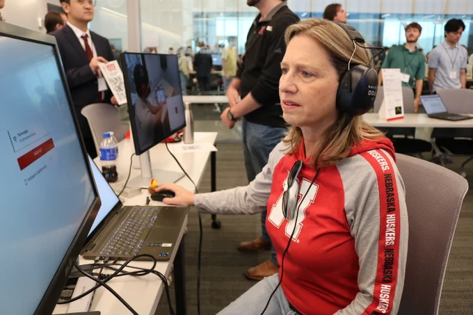 Dr. Ray, a faculty member, tests out hearing equipment used by Dr. Hughes' Cochlear Implant Lab at Senior Design Day. Dr. Ray is wearing a grey and red Nebraska branded hoodie. In the background are students dressed in business attire.