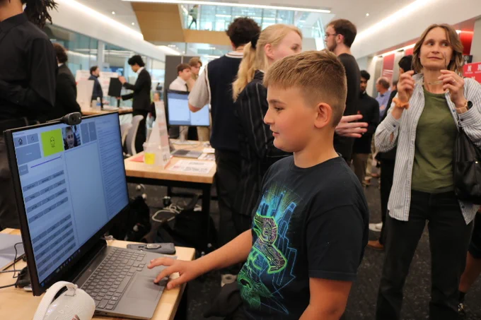 A young boy stands in front of an Augmentative and Alternative Communication device. In the background are students dressed in business attire conversating.