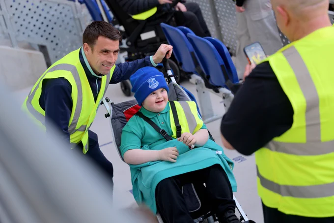 A smiling Seamus Coleman in a high-visibility vest kneels beside a child seated in a wheelchair at a stadium. The child is wearing a blue knit hat, turquoise outfit, and matching safety vest while another person in a safety vest takes their photo with a phone. Blue stadium seats and other people are visible in the background.