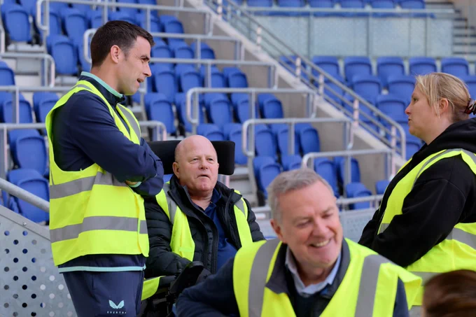 Several people wearing high-visibility safety vests stand and talk in a stadium seating area with rows of bright blue seats in the background. Seamus Coleman stands with his arms crossed beside a man seated in a powered wheelchair, while another man smiles in the foreground and a woman listens on the right.