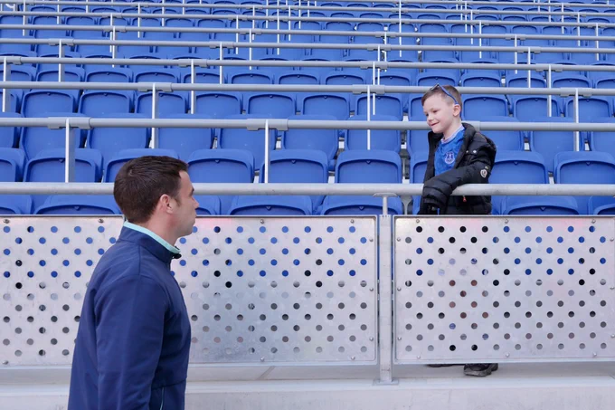 A young boy wearing a blue football shirt and black jacket stands among rows of bright blue stadium seats, smiling down towards Seamus Coleman, who is standing below near a metal barrier.