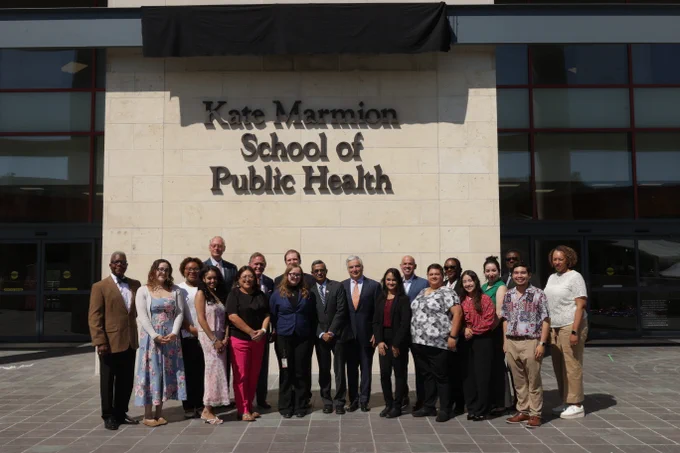 A group of people stands in front of the Kate Marmion School of Public Health building. They are arranged in a line on a paved area, and the building’s name is prominently displayed on the wall behind them.