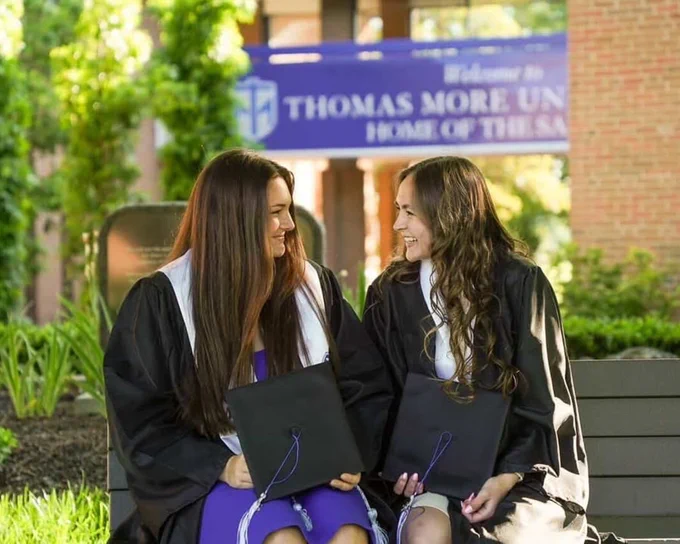 Two graduates sitting on a bench in front of campus wearing graduate gowns and stoles. They are looking at each other while holding a cap and smiling.