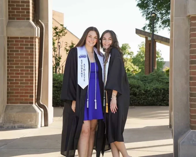 Two graduates in grad attire standing under the bell tower on campus. They are smiling for the camera.