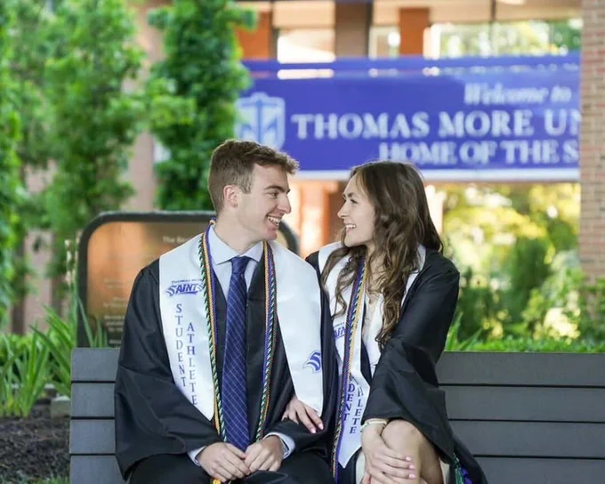 Two graduates sitting on a bench in front of campus wearing graduate gowns and stoles that read "Student Athletes." They are looking at each other and smiling.