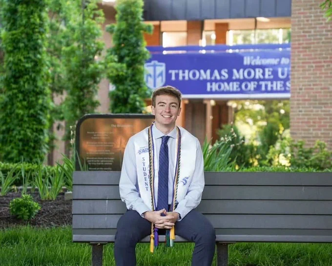 A graduate wearing a stole and cords on a bench in front of campus. He is smiling towards the camera.