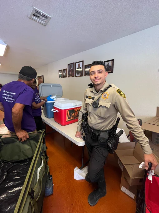 Uniformed LVMPD officer leaning against a table with coolers and boxes in a room with framed photos on the wall.