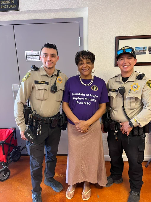 Two uniformed LVMPD officers stand on either side of a woman wearing a purple 'Fountain of Hope Stephen Ministry' shirt inside a community room.