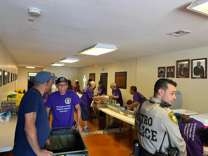 Volunteers in purple shirts and a LVMPD officer packing food and water bottles on tables inside a community center hallway.