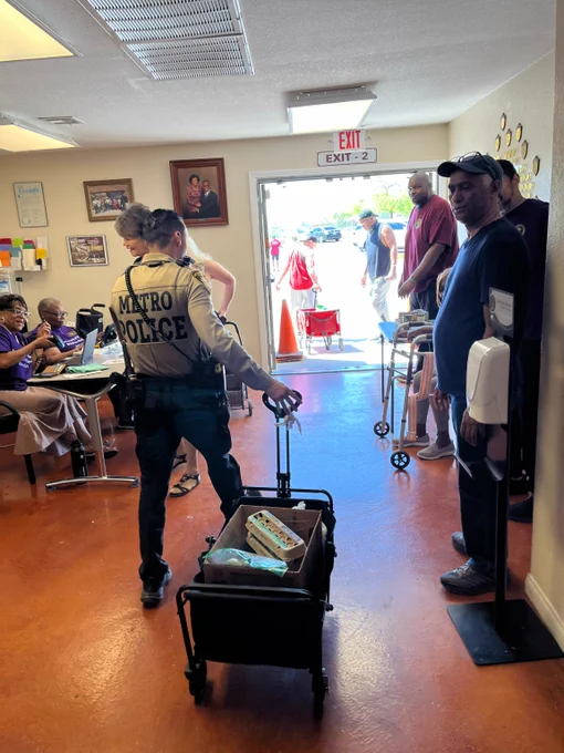 Metro police officer pulling a cart with supplies through a busy community center with people seated and standing nearby.