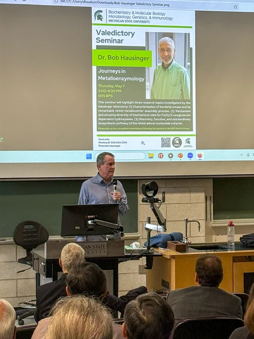 Dr. Scott Mulrooney emcees the event. Behind him on the screen is the seminar flyer. Dr. Hausinger is sitting in the front row, and the back of his head is visible. 