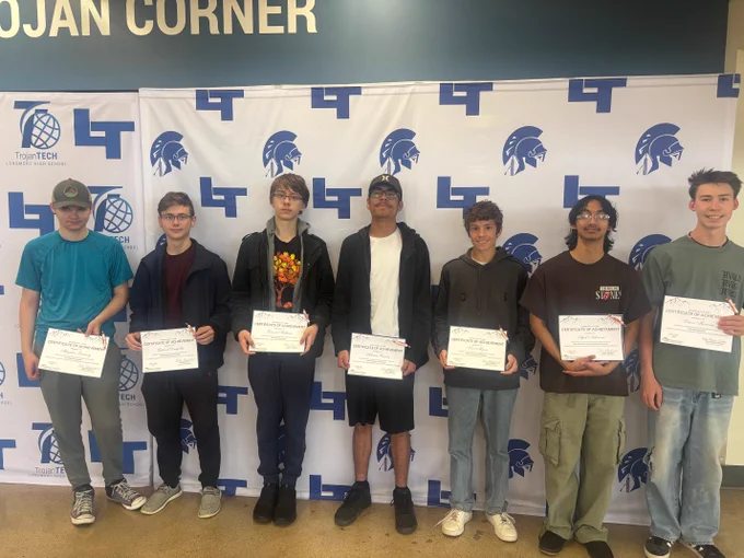 Seven high school students stand in a row in front of a TrojanTech Longmont High School banner beneath a "Trojan Corner" sign, each holding a Certificate of Achievement.